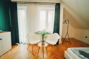 Ein Zimmer mit Holzboden, zwei weiße Stühle, ein kleiner Tisch mit einer Pflanze, ein Bett mit gemusterten Betttextilien und eine Stehlampe.