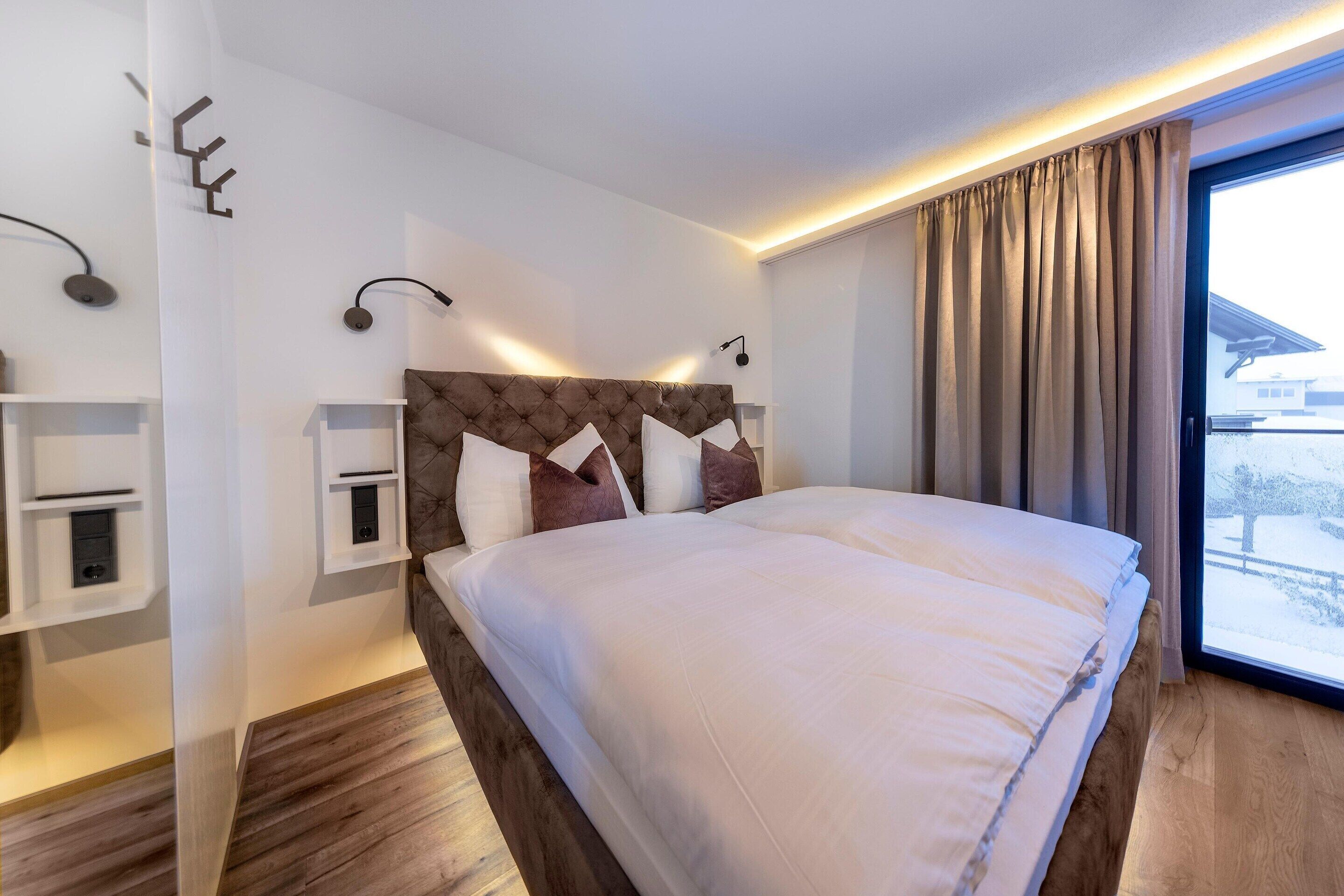 Habitación de hotel moderna con una cama grande, una mesita de noche, un estante en la pared y un ventanal con cortinas.