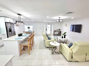 A modern kitchen with a central island, dining area with chairs, a sofa, and a TV mounted on the wall. - Family Pool Retreat - Sleeps 8 - Pet Friendly (Tampa)