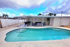 Ein Swimmingpool mit überdachter Terrasse, Grill und Gartenmöbeln.