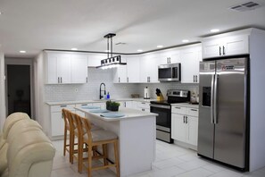 A modern kitchen with white cabinetry, a central island, stainless steel appliances, and a seating area with chairs. - Family Pool Retreat - Sleeps 8 - Pet Friendly (Tampa)