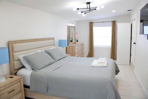 Ein ordentlich bezogenes Bett mit Kopfteil, ein Nachttisch mit Lampe, eine Kommode und ein Fenster mit Vorhängen.