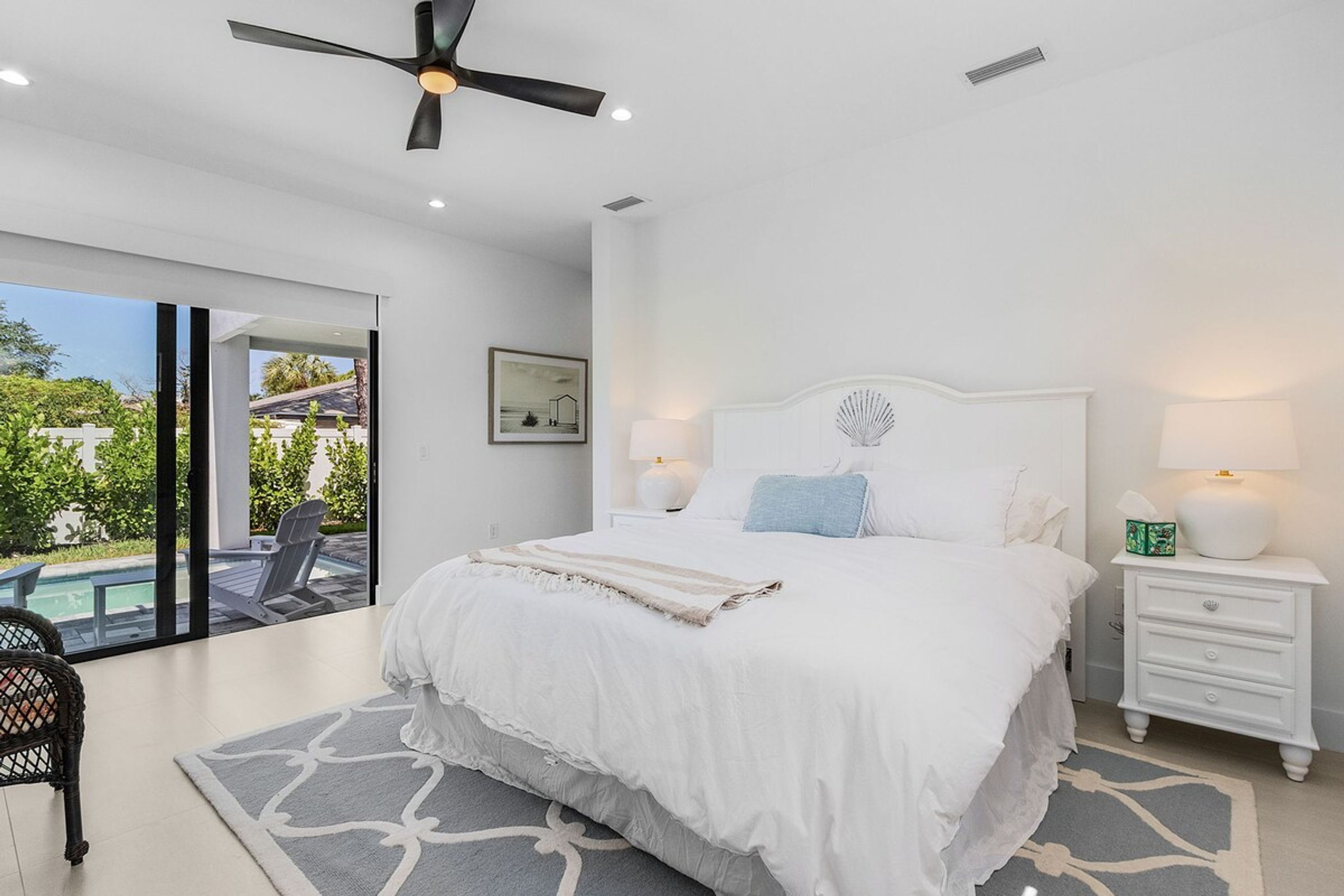 Une chambre avec un grand lit, un ventilateur de plafond, une table de chevet avec une lampe et une vue sur une piscine à l’extérieur.