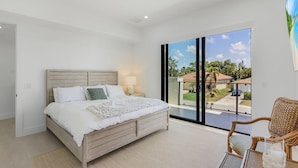 Une chambre avec un grand lit, une table de chevet, une chaise et une vue sur un quartier résidentiel, visible à travers une porte coulissante.