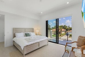 A bedroom with a large bed, a nightstand, a chair, and a view of a residential area through a sliding door. - Modern Naples Beach Home Pool Hot Tub 2 Master Suites (Naples)