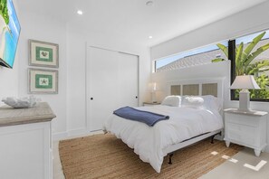A modern bedroom with a bed, bedside tables, a TV, and a window with a view of greenery. - Modern Naples Beach Home Pool Hot Tub 2 Master Suites (Naples)