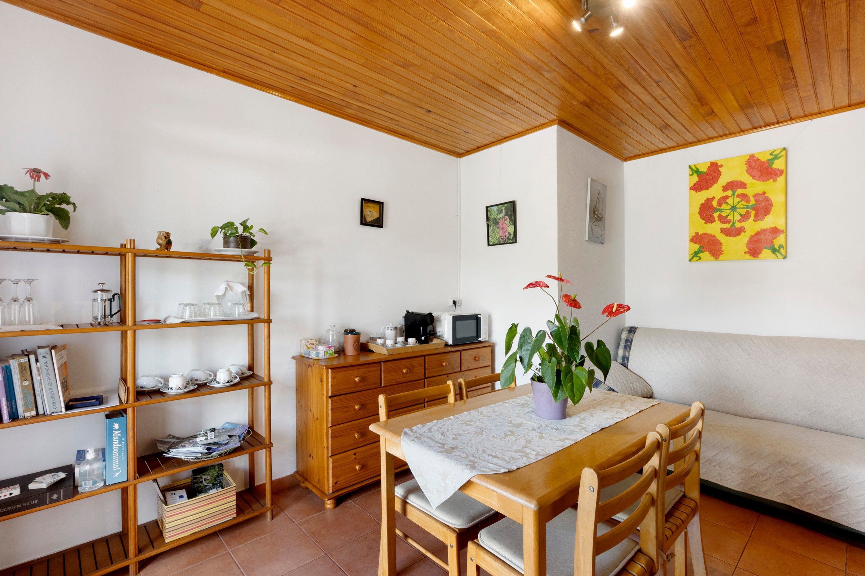 A cozy room with a wooden ceiling, a dining table with chairs, a bookshelf, a microwave, and a potted plant.