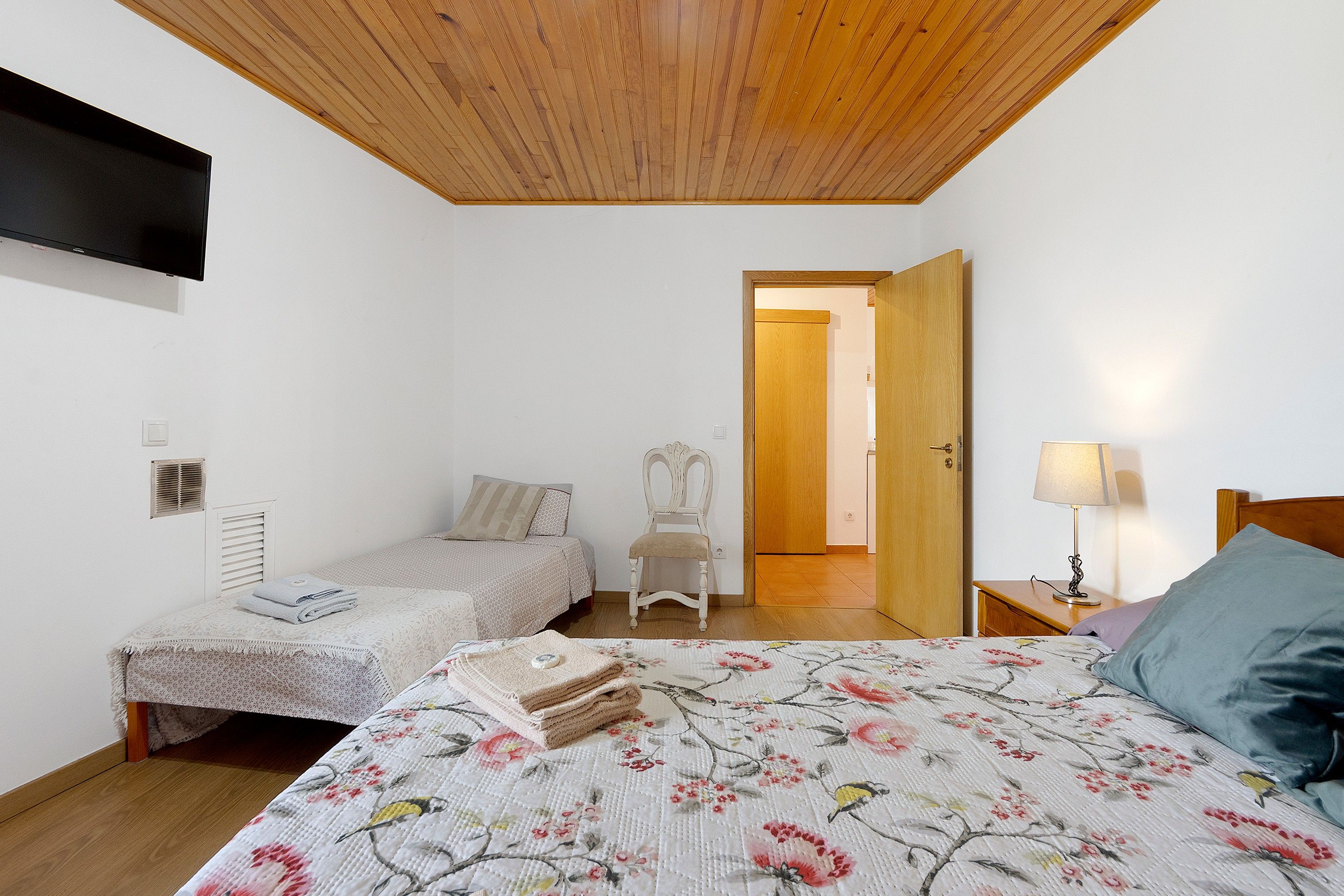 Une chambre avec un plafond en bois, un lit recouvert d’une literie à motifs floraux, une chaise, une table de chevet avec une lampe et une porte donnant sur une autre pièce.