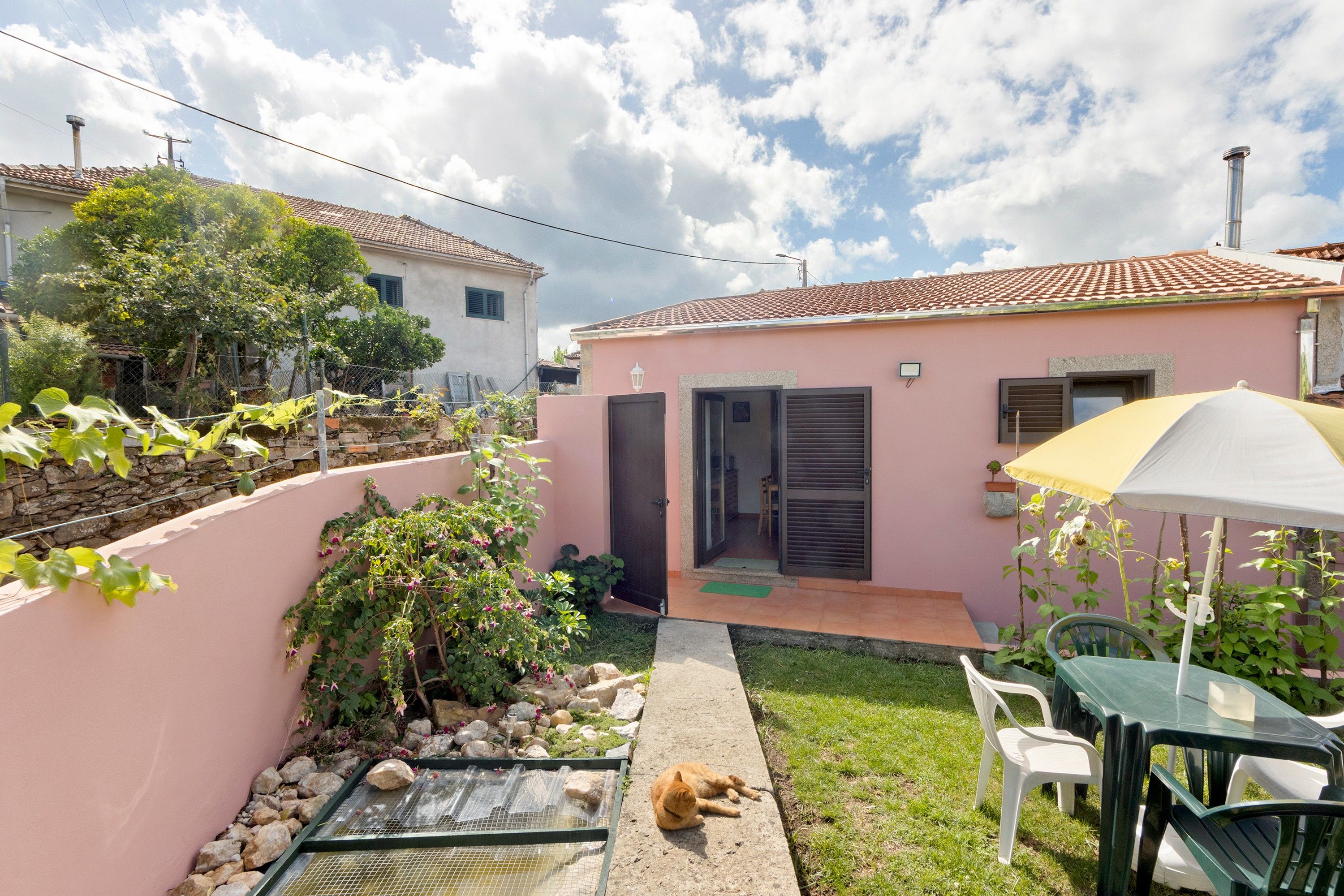 Una propiedad residencial con fachada rosa, techo de tejas, un pequeño jardín y un gato, además de mobiliario de exterior.