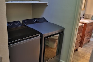 A compact laundry room with a front-loading washer and dryer, a wooden cabinet, and a white countertop. - NEW! Creekside Lounging. Downtown Manitou Springs. 2 BR/2 BA. A/C. New Hot Tub! (Manitou Springs)