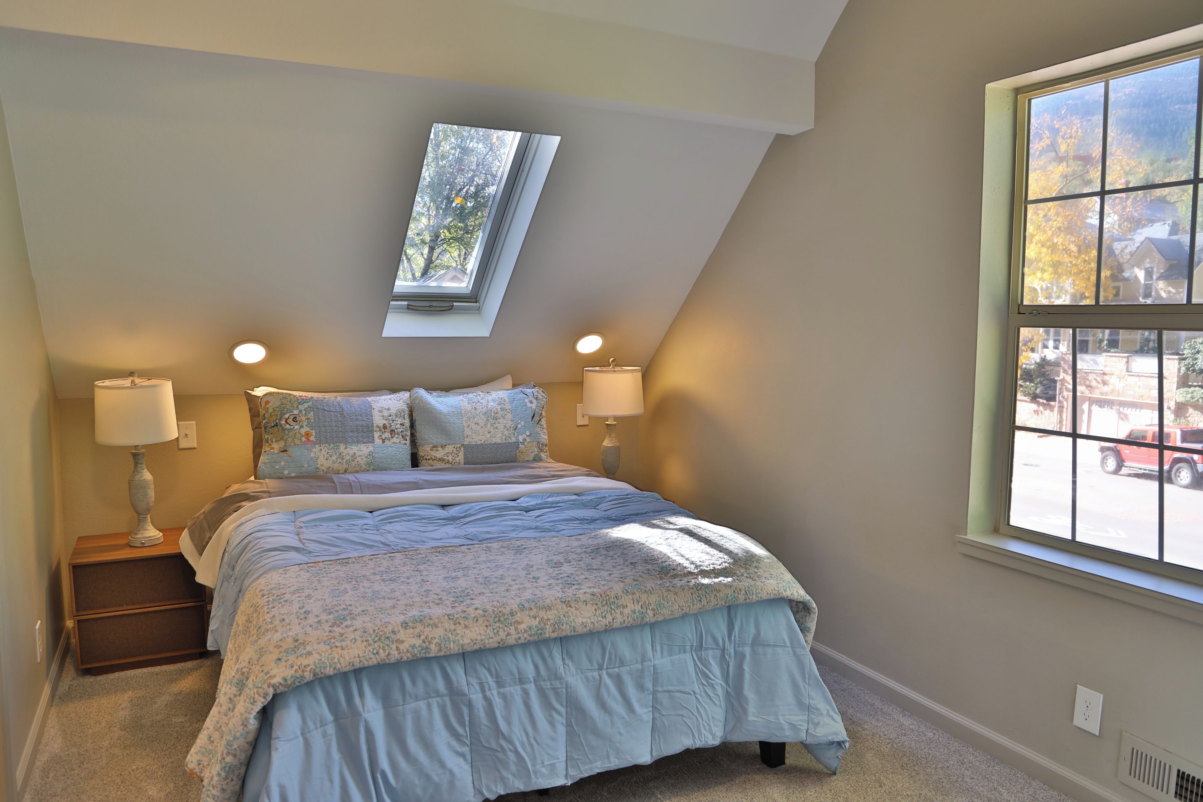 Ein Schlafzimmer mit einem Oberlicht, zwei Nachttischlampen, einer blumigen Tagesdecke und Blick ins Freie durch ein großes Fenster.