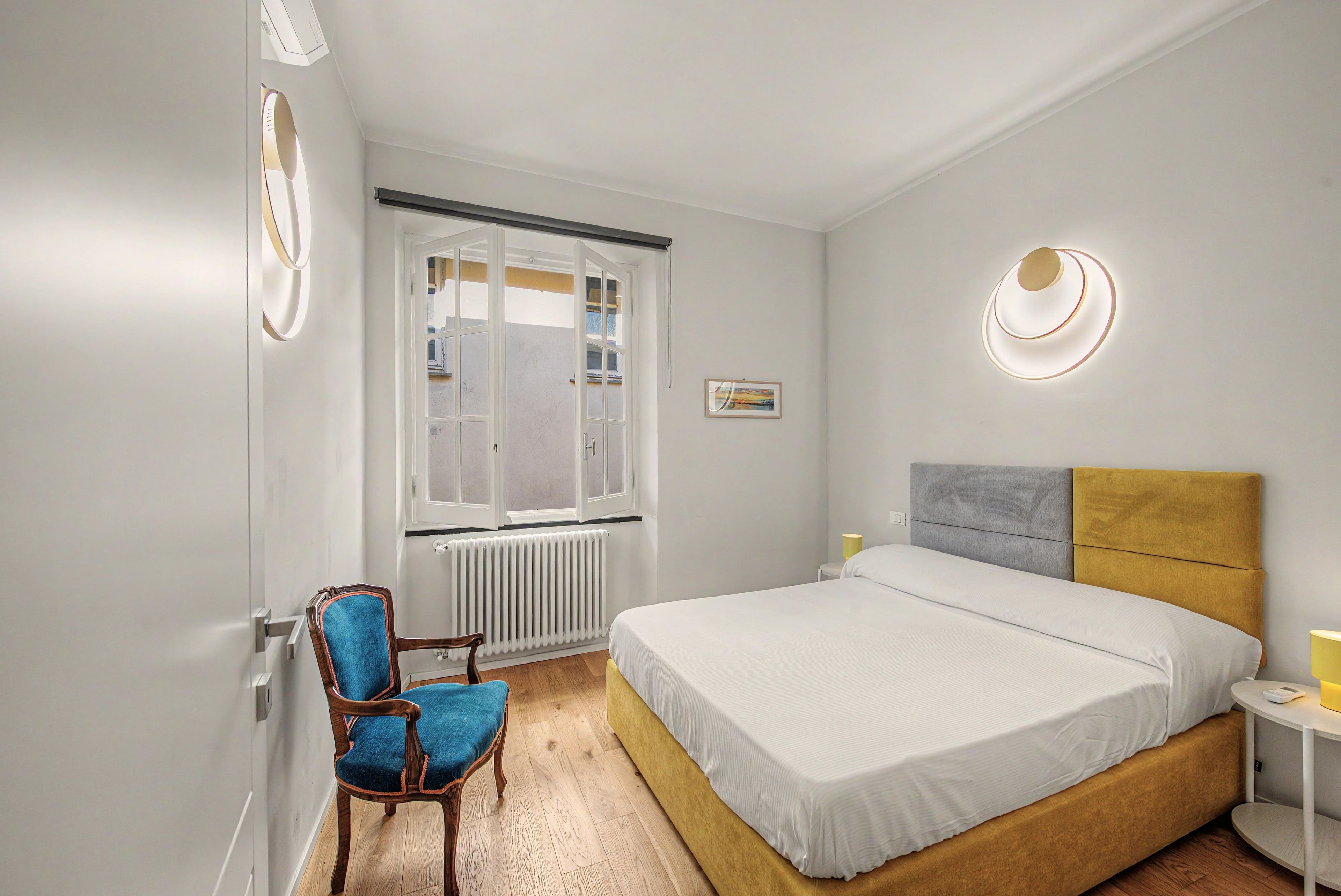 Ein Schlafzimmer mit Bett, Sessel, Heizkörper, Fenster und einer Wandleuchte.