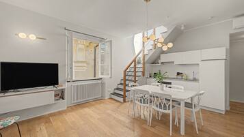 Eine moderne Küche mit weißen Schränken, eine Essecke mit weißem Tisch und Stühlen, ein integrierter Fernseher und eine Treppe, die in ein Obergeschoss führt.