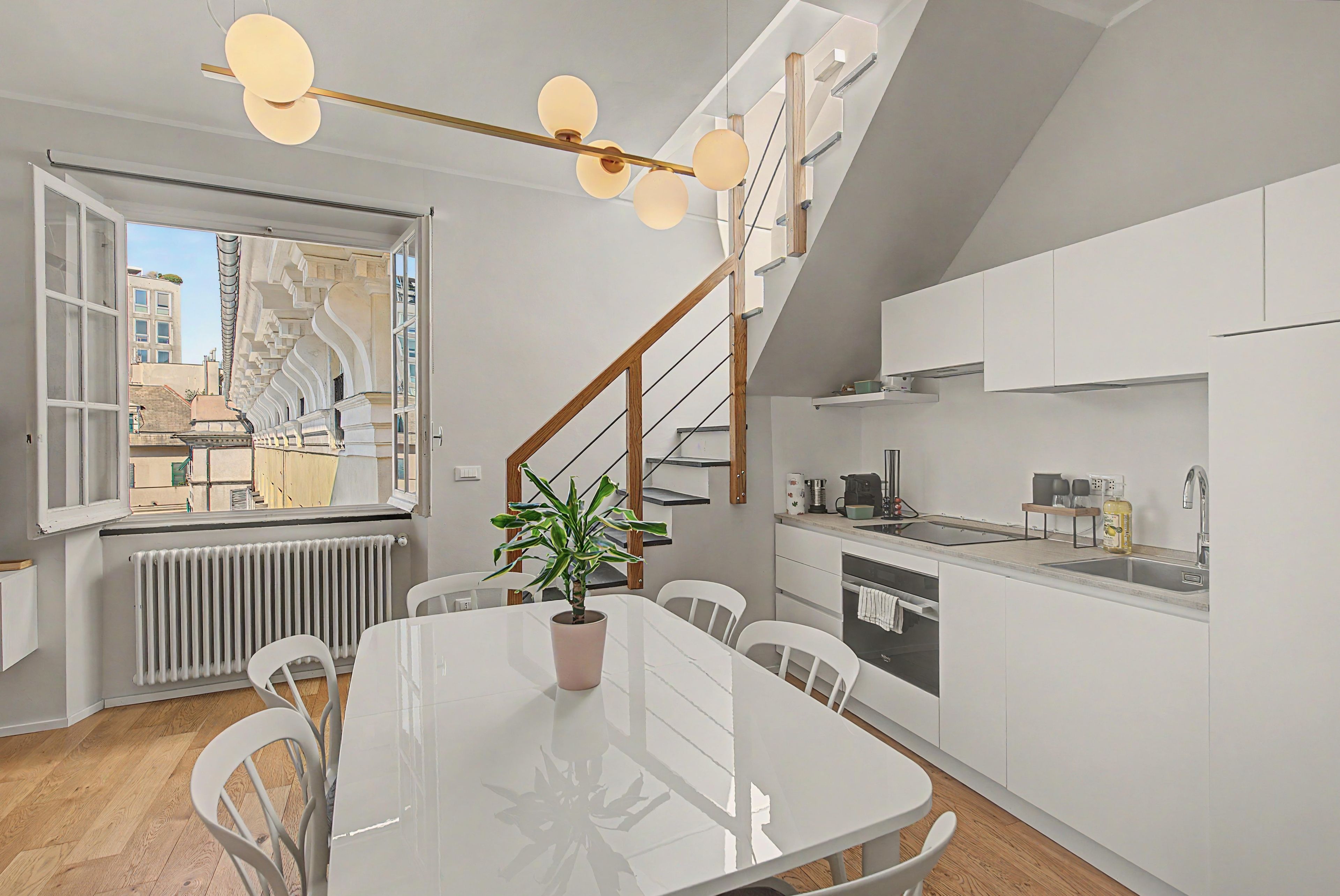 A modern kitchen with white cabinetry, a dining table with chairs, a potted plant, and a staircase with a wooden railing.