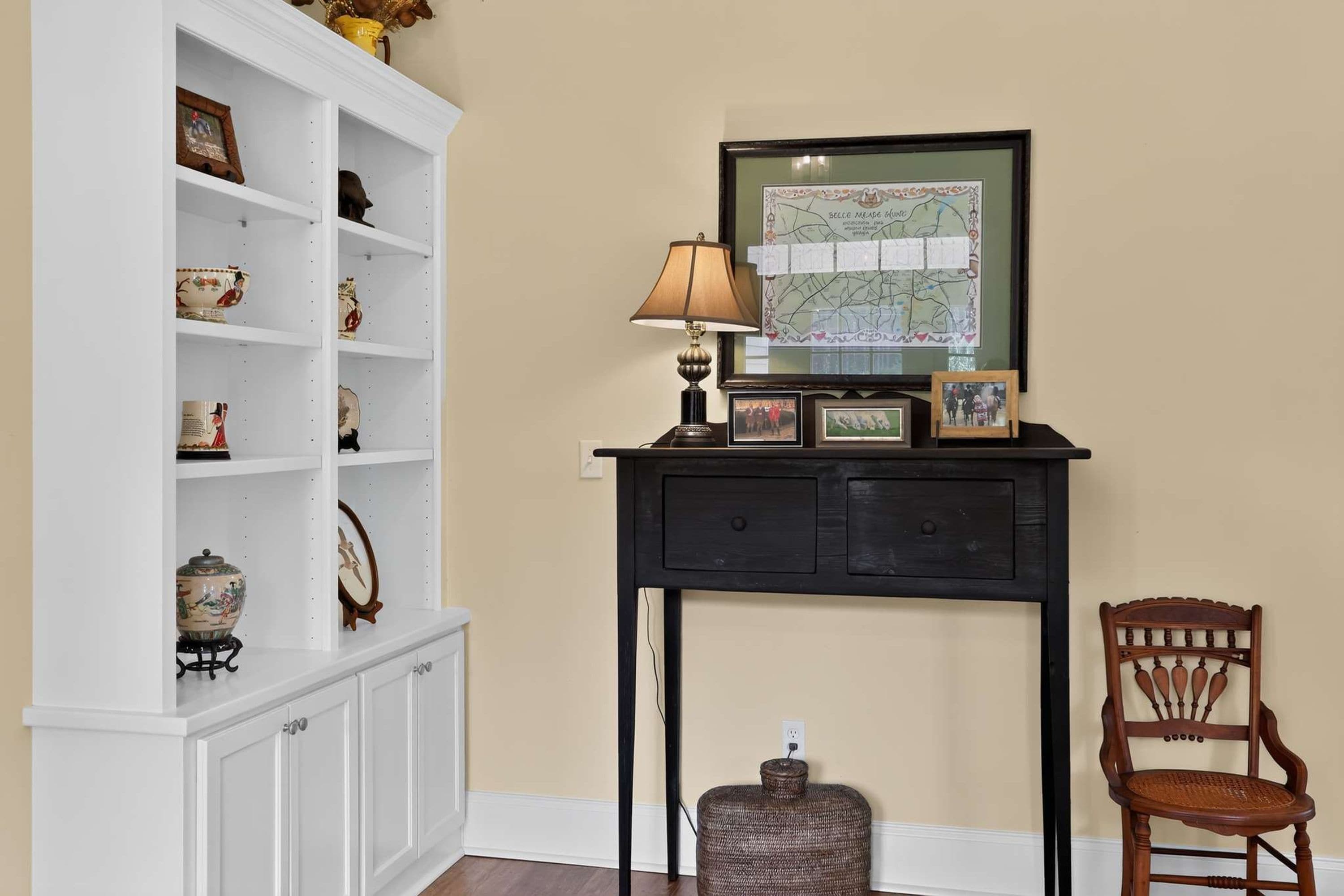 A room with a white shelving unit, a black console table with a lamp, a framed map, and a wooden chair.