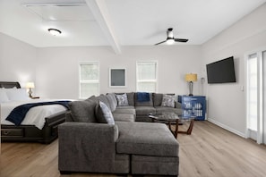 Un salon moderne avec un canapé d’angle, une table basse en verre, un lit avec une tête de lit et un ventilateur de plafond.