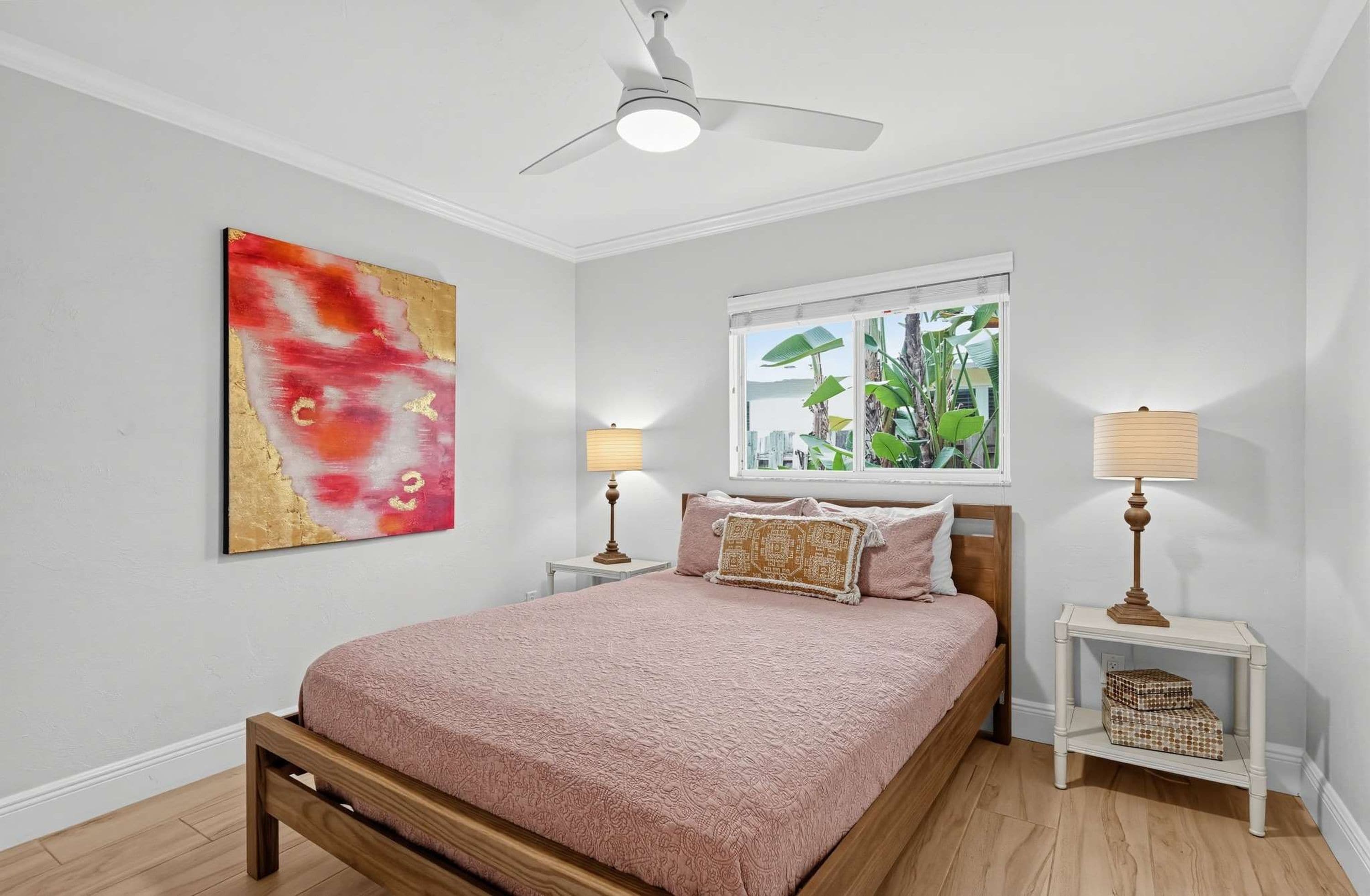 A bedroom with a bed, two bedside tables, a ceiling fan, a window with a view, and a wall-mounted abstract painting.