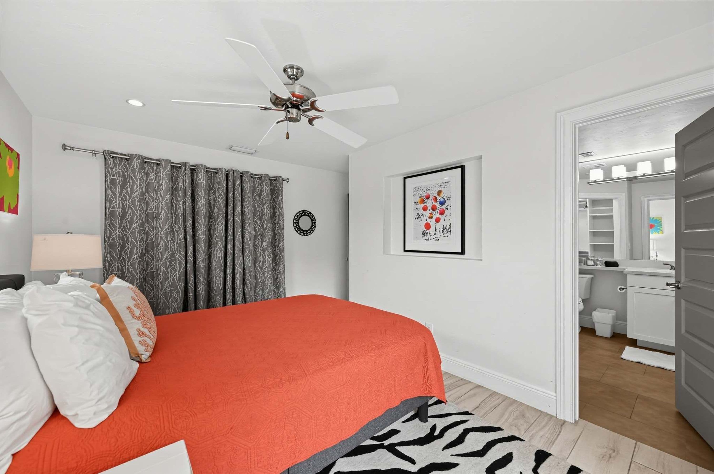 A bedroom with a bed, a ceiling fan, a bathroom visible through an open door, and a framed picture on the wall.
