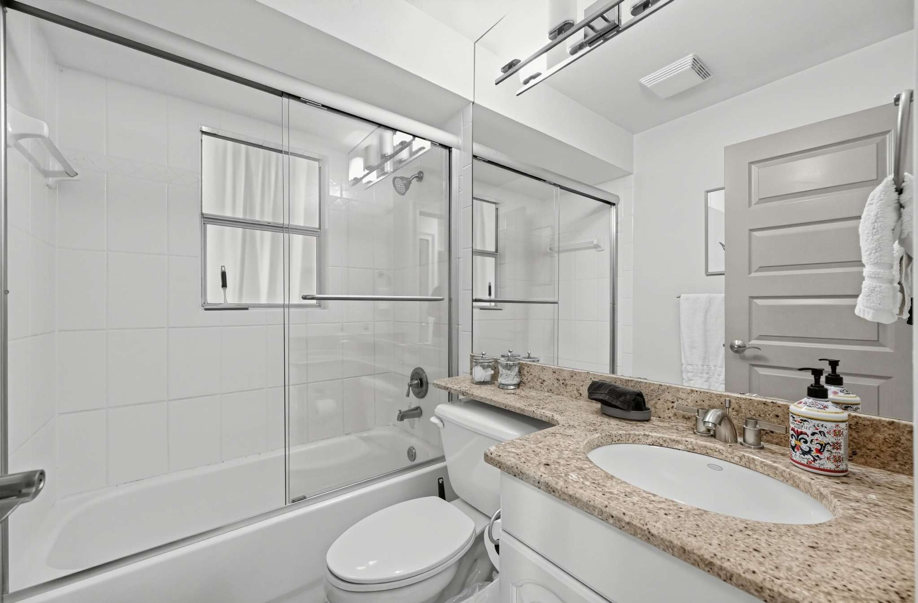 A modern bathroom with a glass-enclosed shower, a white toilet, a sink with a granite countertop, and a large mirror.