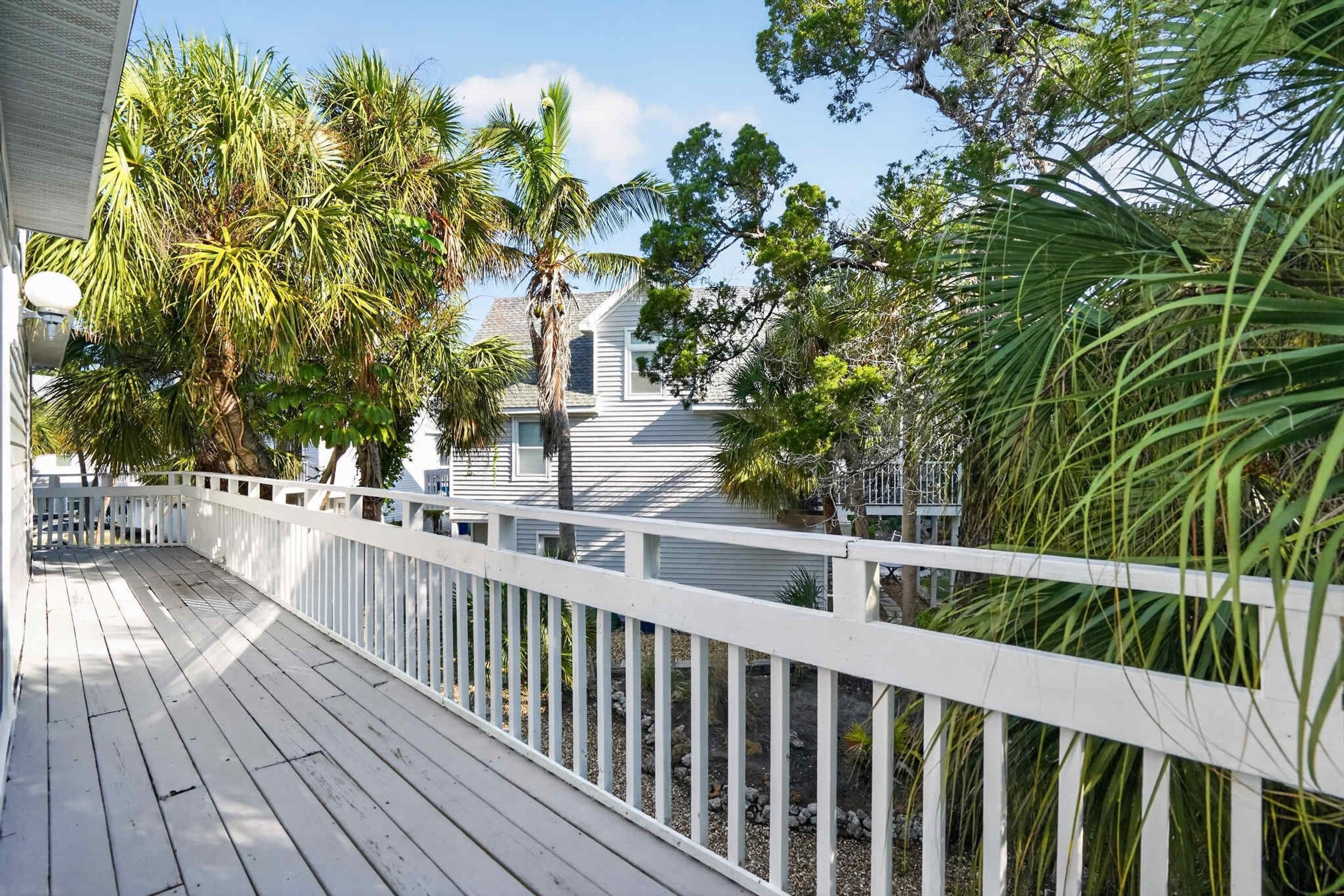 A wooden deck with a white railing, surrounded by lush greenery and a house in the background.