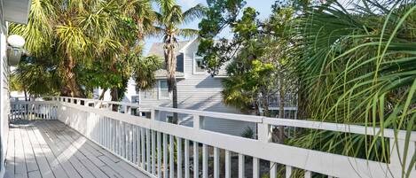 A wooden deck with a white railing, surrounded by lush greenery and a house in the background.