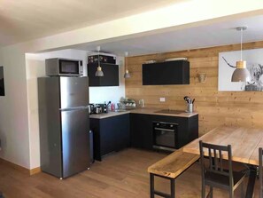 A modern kitchen with a built-in microwave, refrigerator, and oven. There is a wooden dining table with chairs and a hanging pendant light. - Off-Piste | At the Foot of the Slopes in Val Louron (Génos)