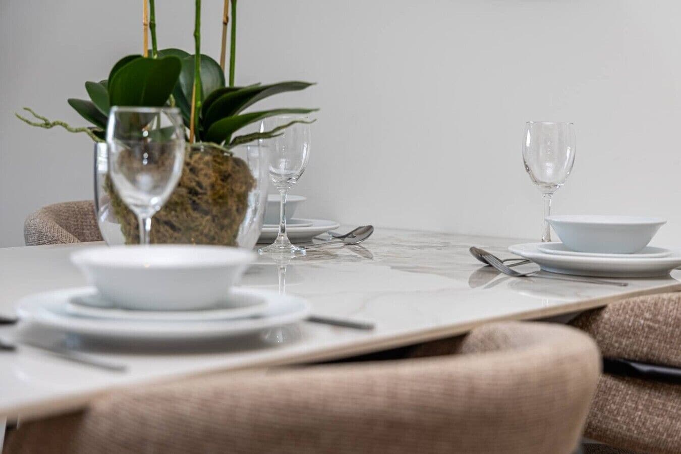 A dining table set with white plates, bowls, and cutlery, accompanied by two wine glasses and a potted plant.