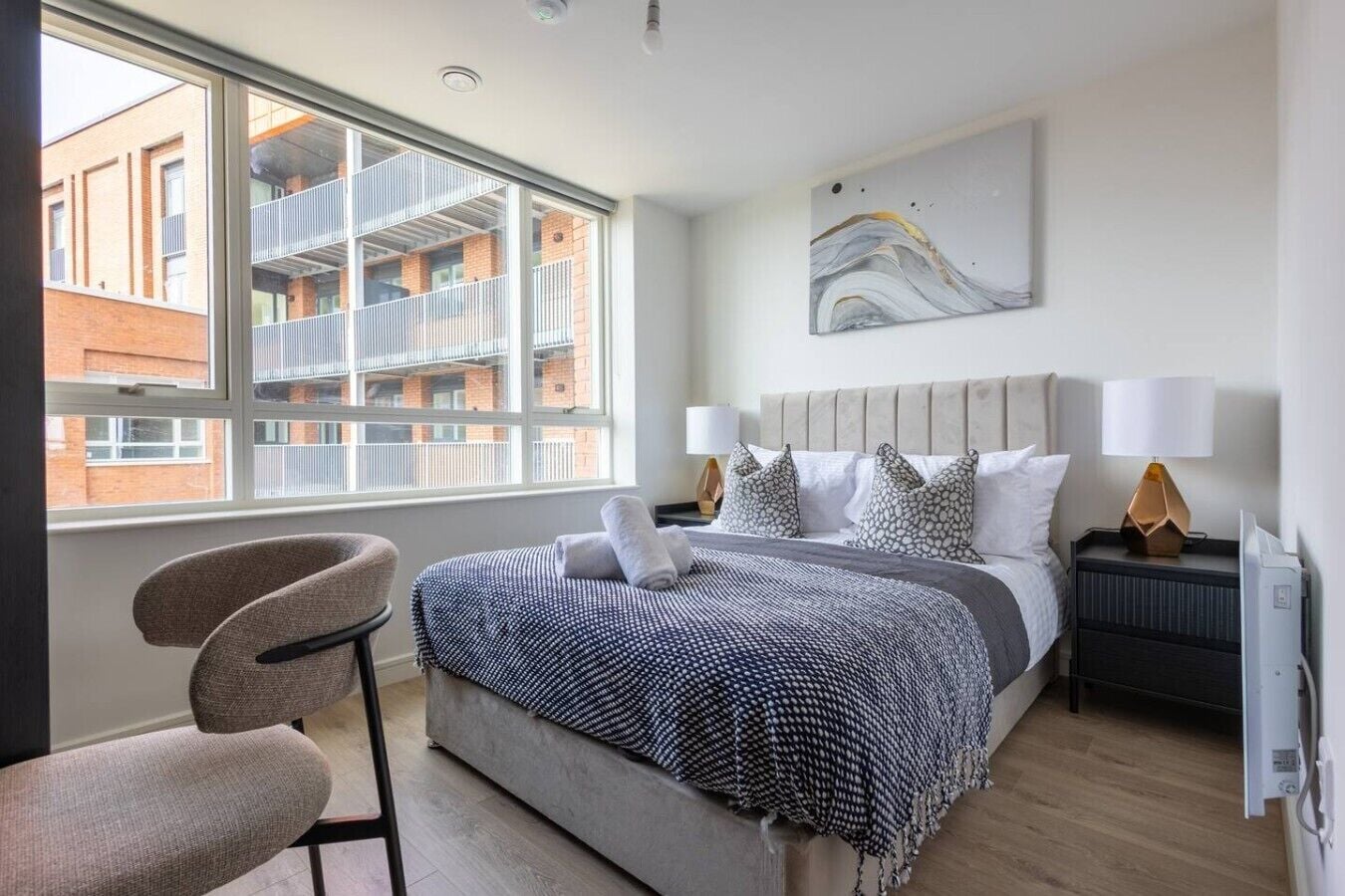 A hotel room with a bed, a chair, a window with a view of buildings, and a painting on the wall.