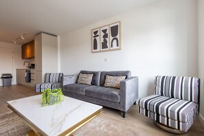 Un salon avec un canapé gris, des fauteuils à rayures, une table basse en marbre et, au fond, une cuisine.