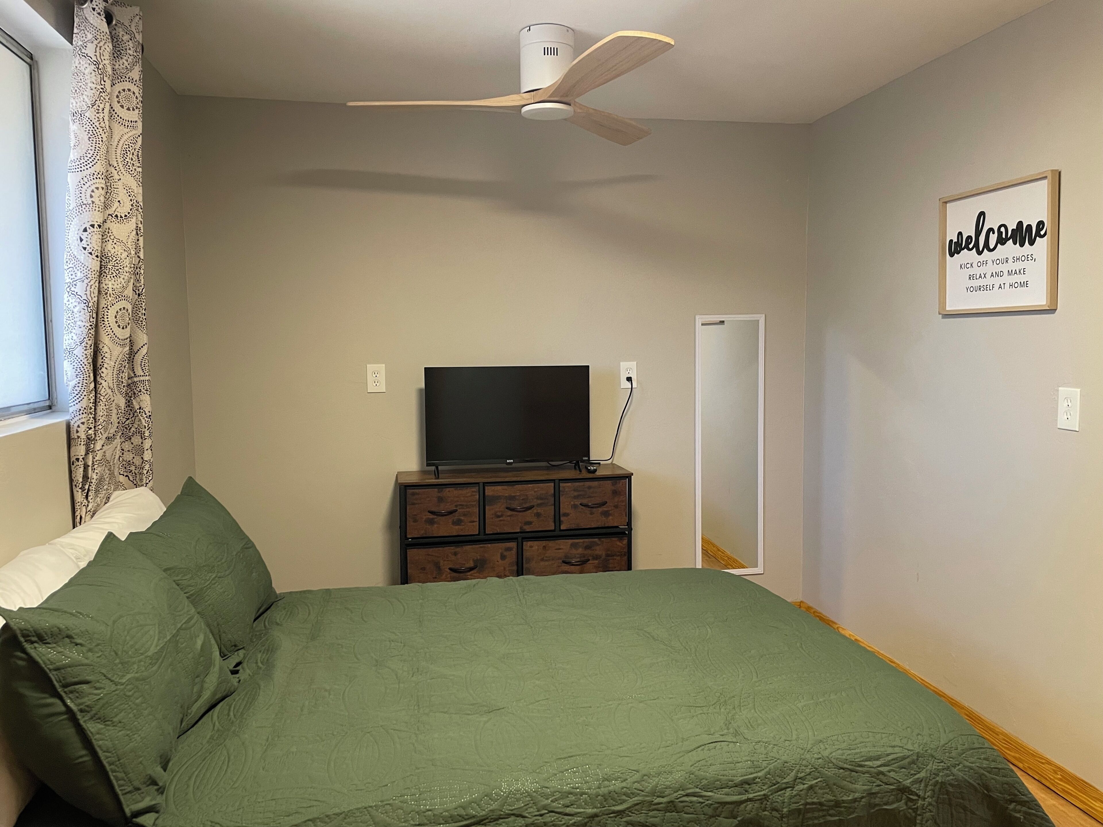 Un dormitorio con cama, cómoda, televisor, ventilador de techo y un cartel de bienvenida.