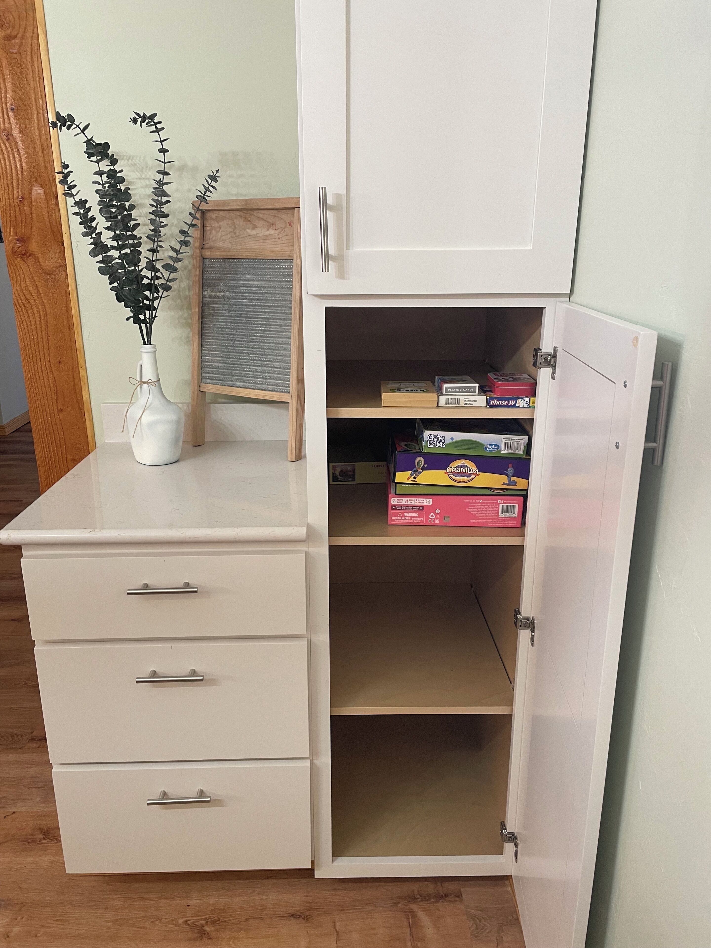 A white cabinet with a drawer and a shelf, containing a few items including a box of playing cards and a box of cereal.
