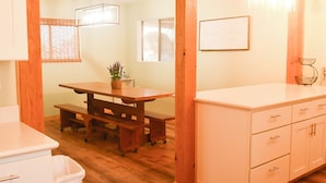 Een kamer met een houten eettafel, bankjes, een wit kastje met lades en een bord met een raam op de muur.