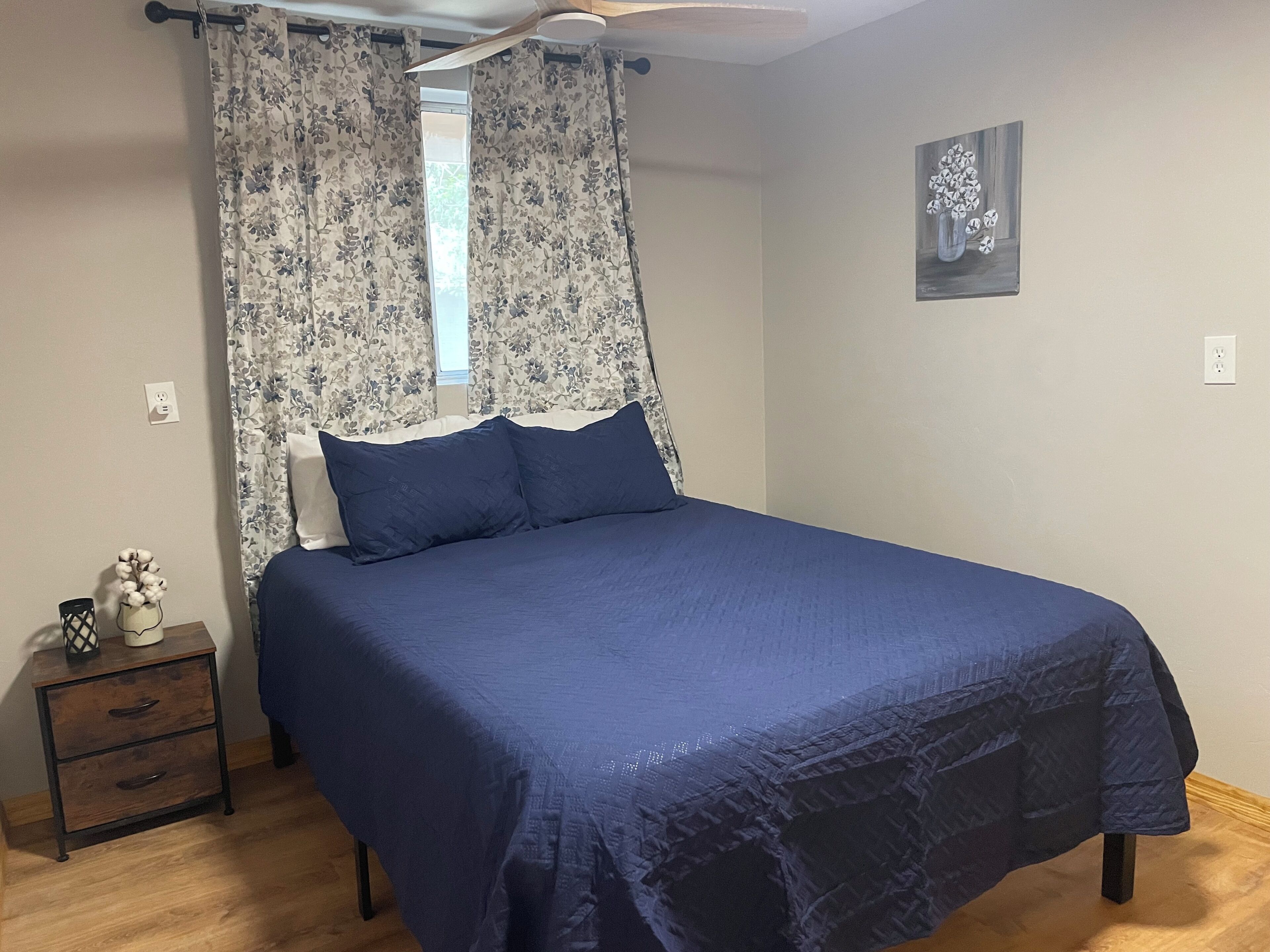 Un dormitorio con cama, mesita de noche, un florero y un cuadro en la pared.