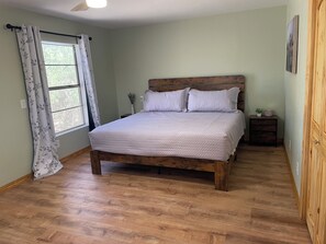 A bedroom with a wooden bed, a window with curtains, a nightstand, and a wall-mounted picture.