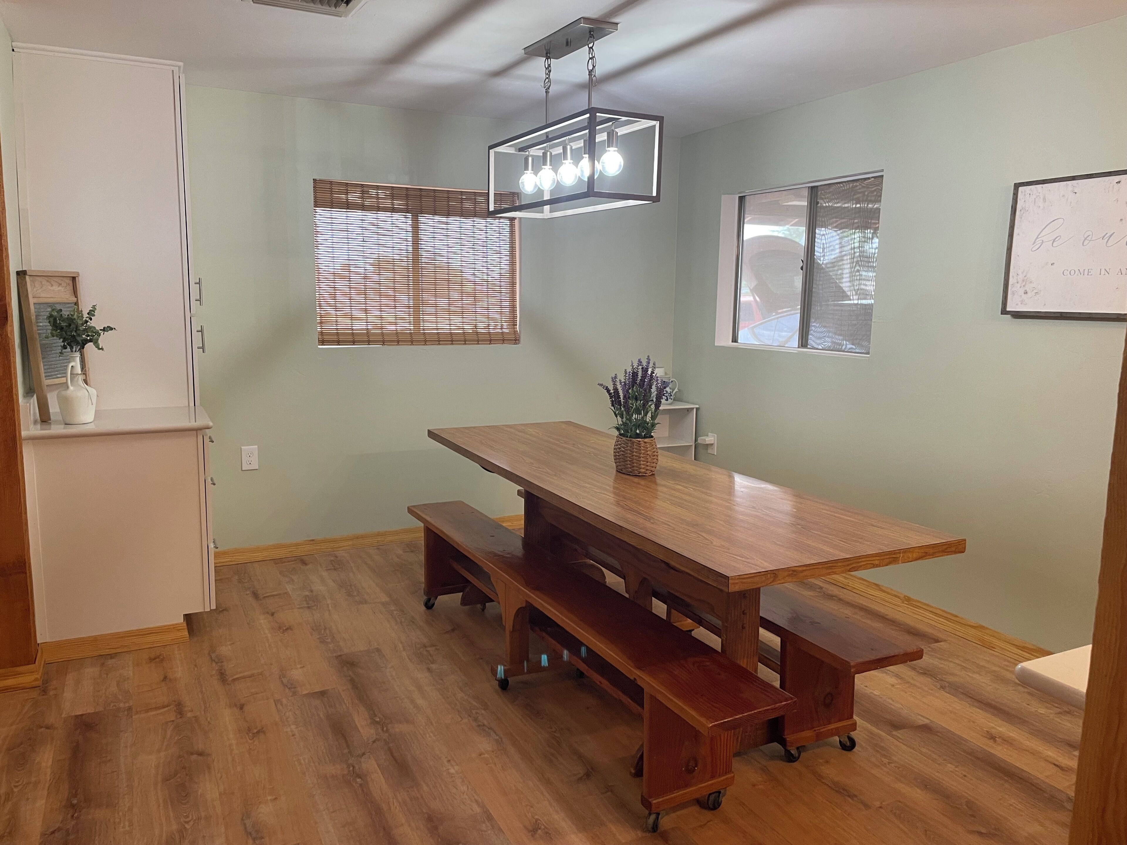 Un comedor con mesa de madera y bancos, lámpara colgante y ventana con persianas.