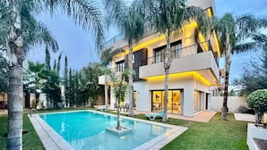 A modern multi-story building with a swimming pool, surrounded by palm trees and well-maintained grass. - PREMIUM EXPERIENCE - MODERN VILLA WITH EXCEPTIONAL COMFORT (مراكش)