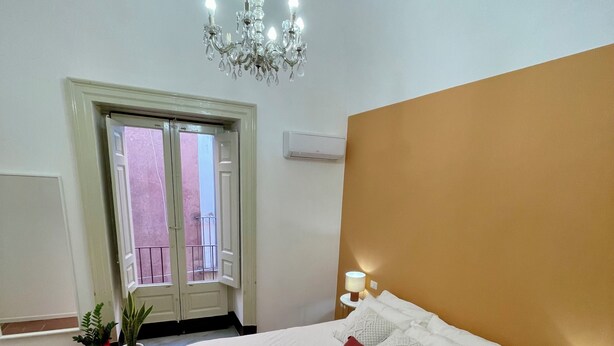 Un dormitorio con cama, candelabro, mesita de noche con lámpara, planta en maceta y vista al balcón exterior.