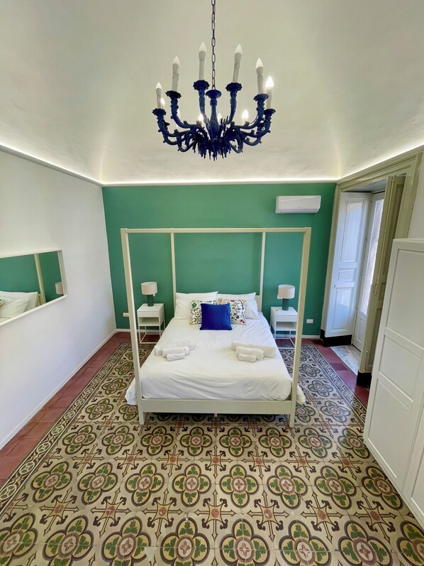 Un lit à baldaquin avec du linge de lit blanc, un oreiller bleu et des serviettes blanches posées sur le sol. Un lustre est suspendu au-dessus du lit, et la pièce est décorée de murs verts et de carreaux de sol à motifs.