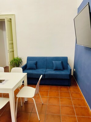 A room with a blue sofa, a white table, and a white chair. There is a plant on the floor and a door in the background. - Fil's House—located in the historic district, close from the botanical garden (Catania)