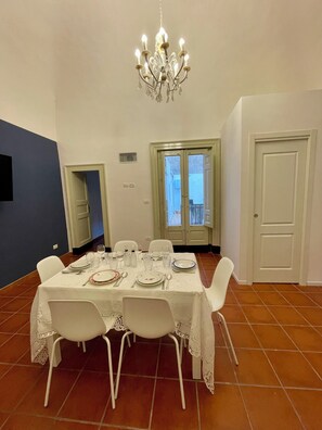 Une salle à manger agrémentée d’un lustre, avec une table blanche dressée pour quatre personnes et une vue sur l’extérieur grâce à une porte vitrée.