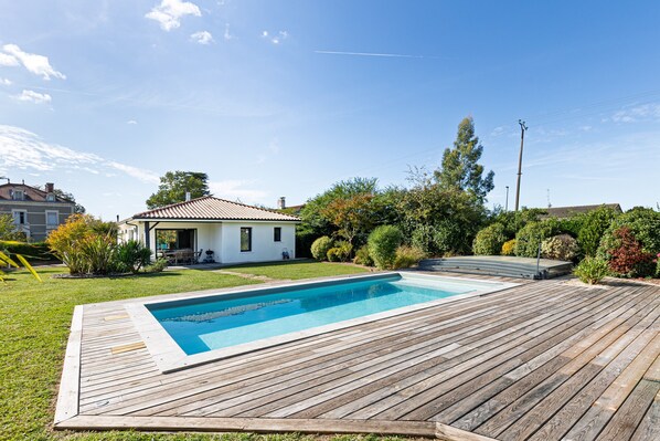 A swimming pool with a wooden deck, surrounded by a well-maintained lawn and various shrubs.