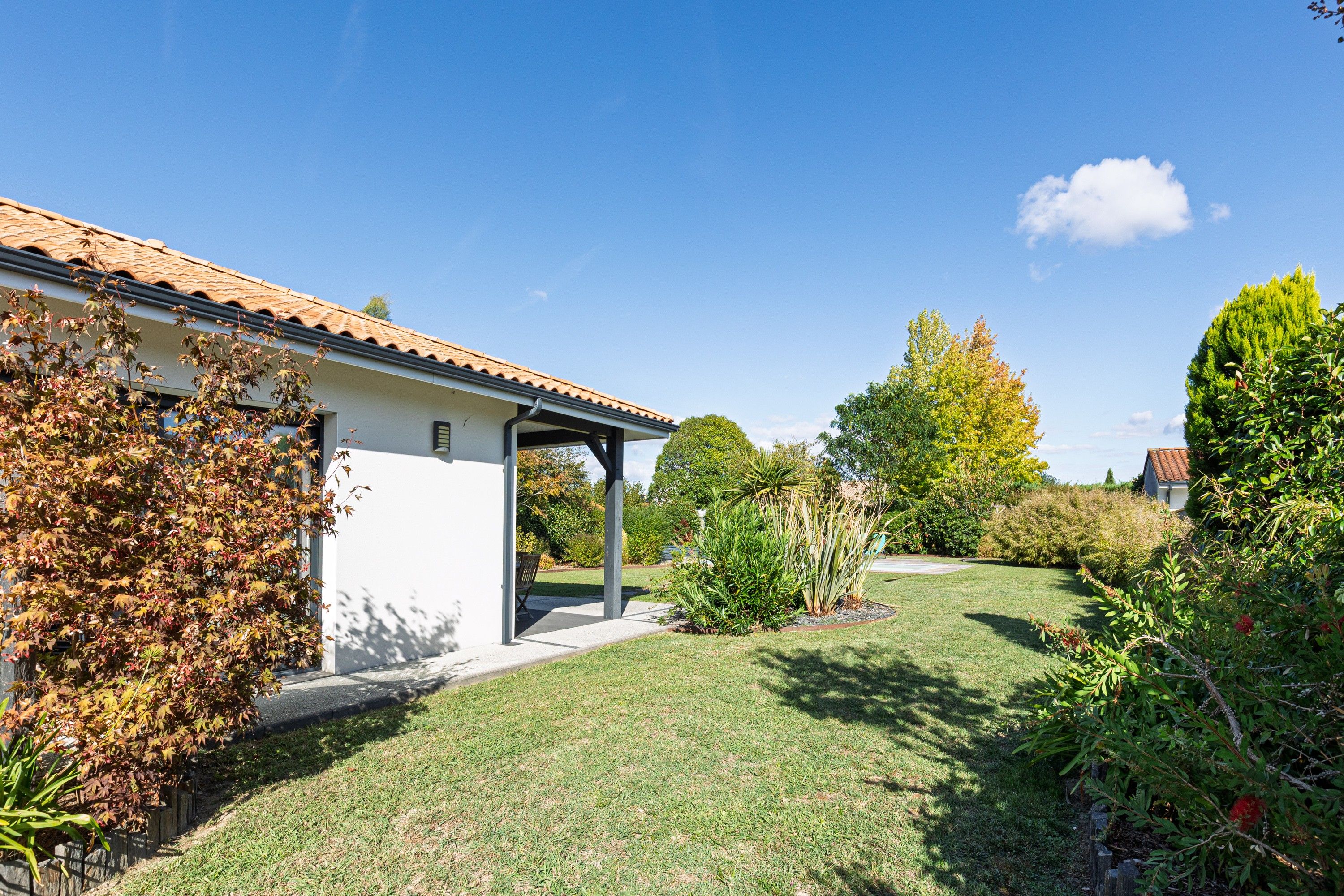 Una casa amb terrassa coberta, gespa i diverses plantes i arbustos.
