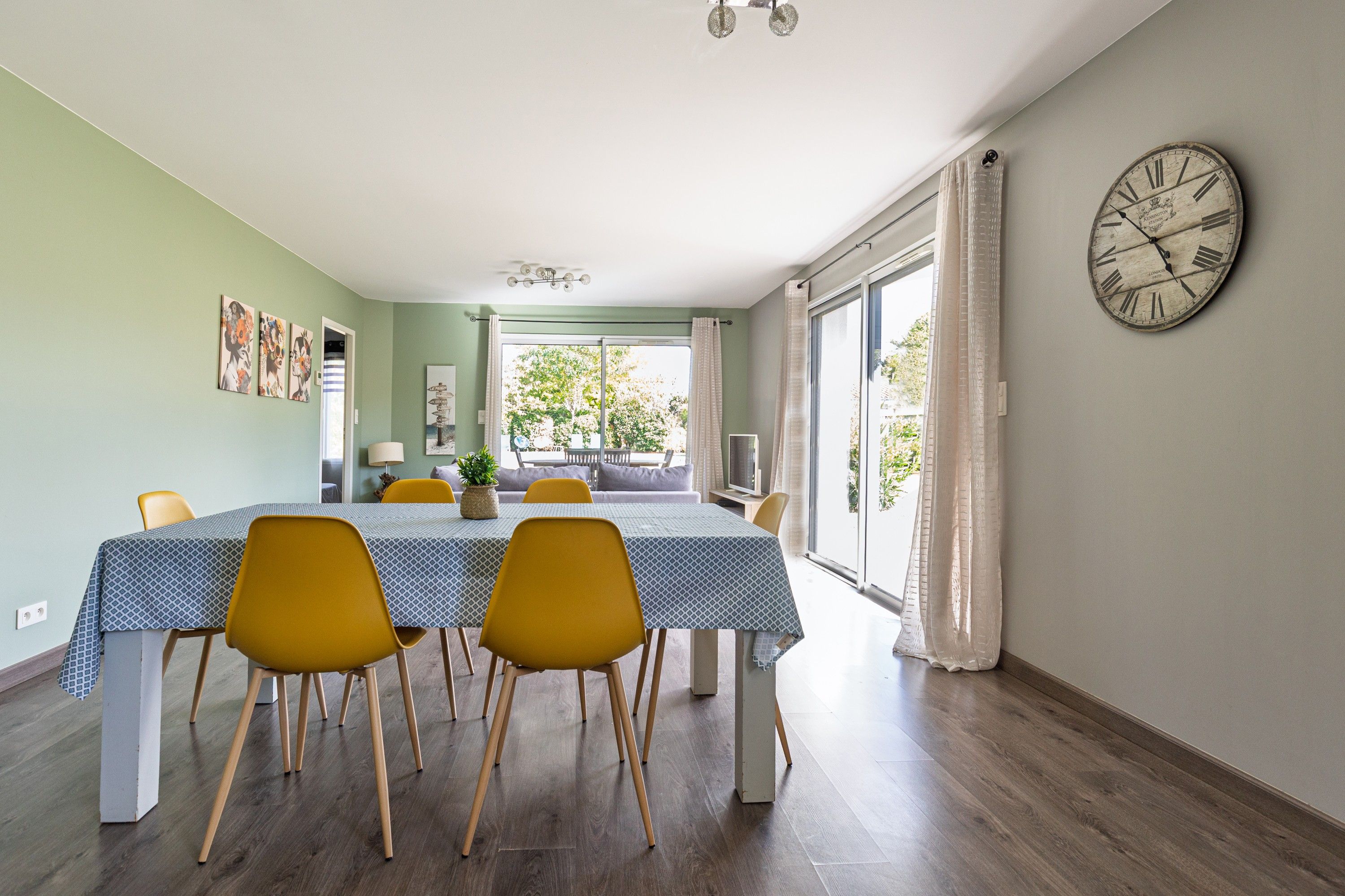 Un coin repas avec une table dressée pour quatre personnes, des chaises, une horloge au mur et une vue à travers la porte vitrée.