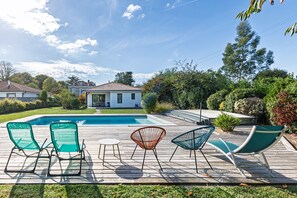 A swimming pool with a wooden deck, surrounded by greenery and a house in the background.