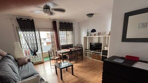 Uma sala de estar com sofá, mesa de jantar e cadeiras, TV sobre um suporte e ventilador de teto.