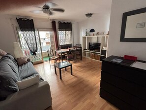 Una sala de estar con un sofá, una mesa de comedor con sillas, un televisor sobre un mueble bajo y un ventilador de techo.
