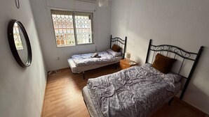 Quarto com duas camas de solteiro, mesa de cabeceira iluminada por um abajur, janela com vista para prédios e ventilador de teto.