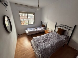 Un dormitorio con dos camas individuales, una mesita de noche con lámpara, una ventana con vistas a edificios y un ventilador de techo.