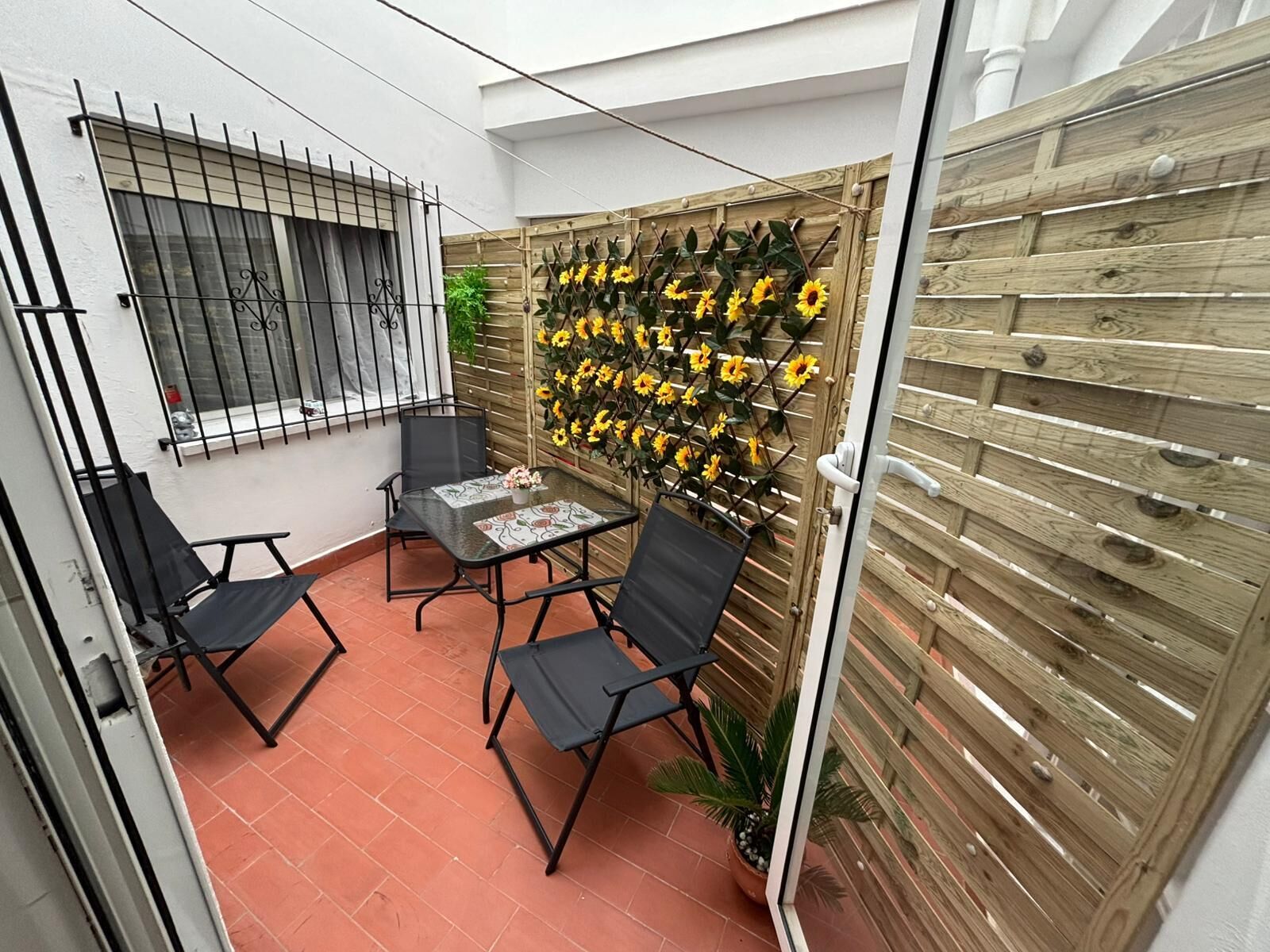 Eine Terrasse mit Tisch und Stühlen, ein Holzzaun mit Sonnenblumen und ein Fenster mit Metallgitter.