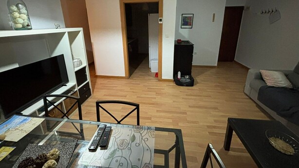 Uma sala de estar com sofá, televisão, mesa de jantar com cadeiras, uma mesa de vidro com controles remotos, ventilador de teto e um corredor que dá para outro cômodo.